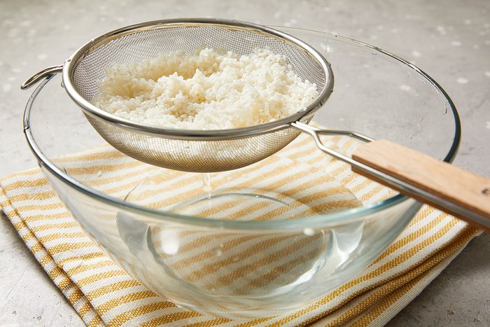 Closeup shot of a metal strainer holding kefir grains; set over a clear glass bowl to drain the liquid; both placed on a yellow-and-white striped cloth on a light-colored surface.