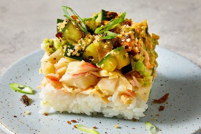 Closeup shot of a single square serving of Sushi Bake; layered with fluffy white rice, shredded imitation crab, creamy toppings, diced cucumber, toasted breadcrumbs, and sliced green onions; presented on a light blue speckled plate;