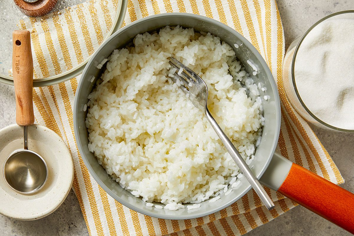 Overhead shot of a pot of cooked white rice with a fork resting inside; set on a yellow-striped towel; nearby sit a metal measuring spoon on a small dish, a bowl of sugar, and a partially visible glass lid.