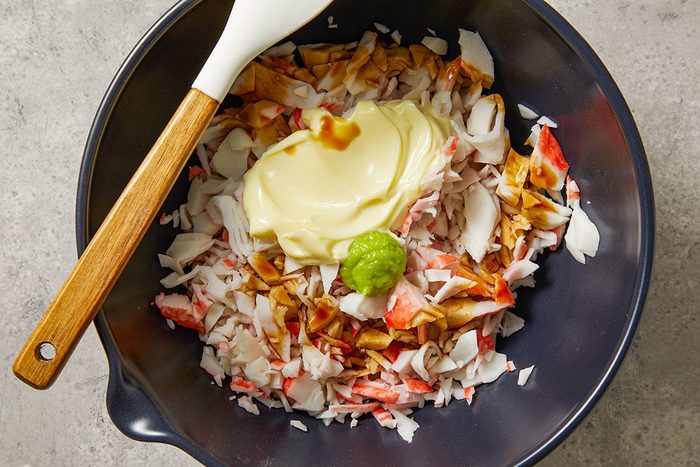 Overhead shot of a black mixing bowl containing chopped imitation crab; a dollop of mayonnaise; a small scoop of green wasabi; and a splash of soy sauce; a white spatula rests on the edge of the bowl;