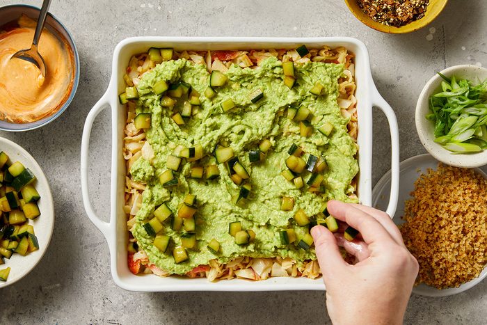 Overhead shot of a hand sprinkling diced cucumber over a casserole topped with guacamole in a white baking dish; surrounding bowls hold sauce, sliced green onions, seasonings, and breadcrumbs on a light countertop;