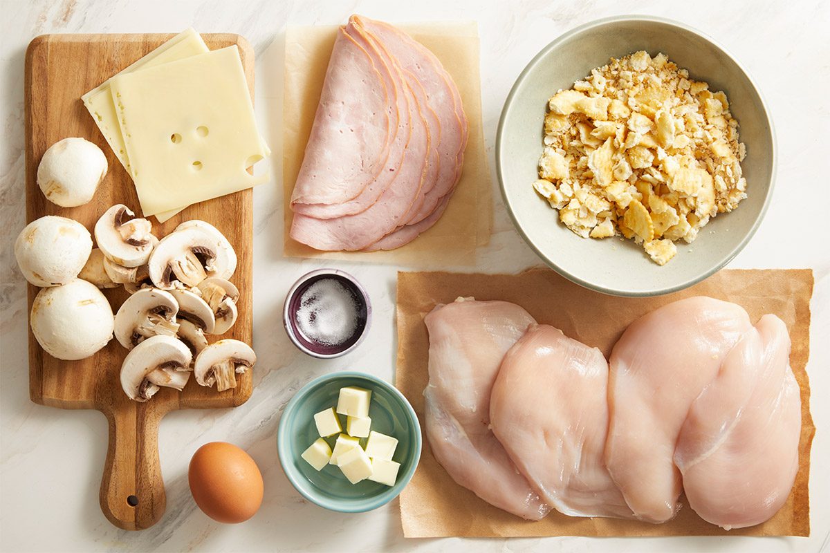 Overhead shot of ingredients, raw chicken breasts, Swiss cheese, mushrooms, ham, egg, and crushed crackers arranged on a light wooden surface.