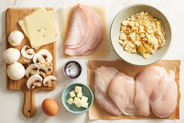 Overhead shot of ingredients, raw chicken breasts, Swiss cheese, mushrooms, ham, egg, and crushed crackers arranged on a light wooden surface.