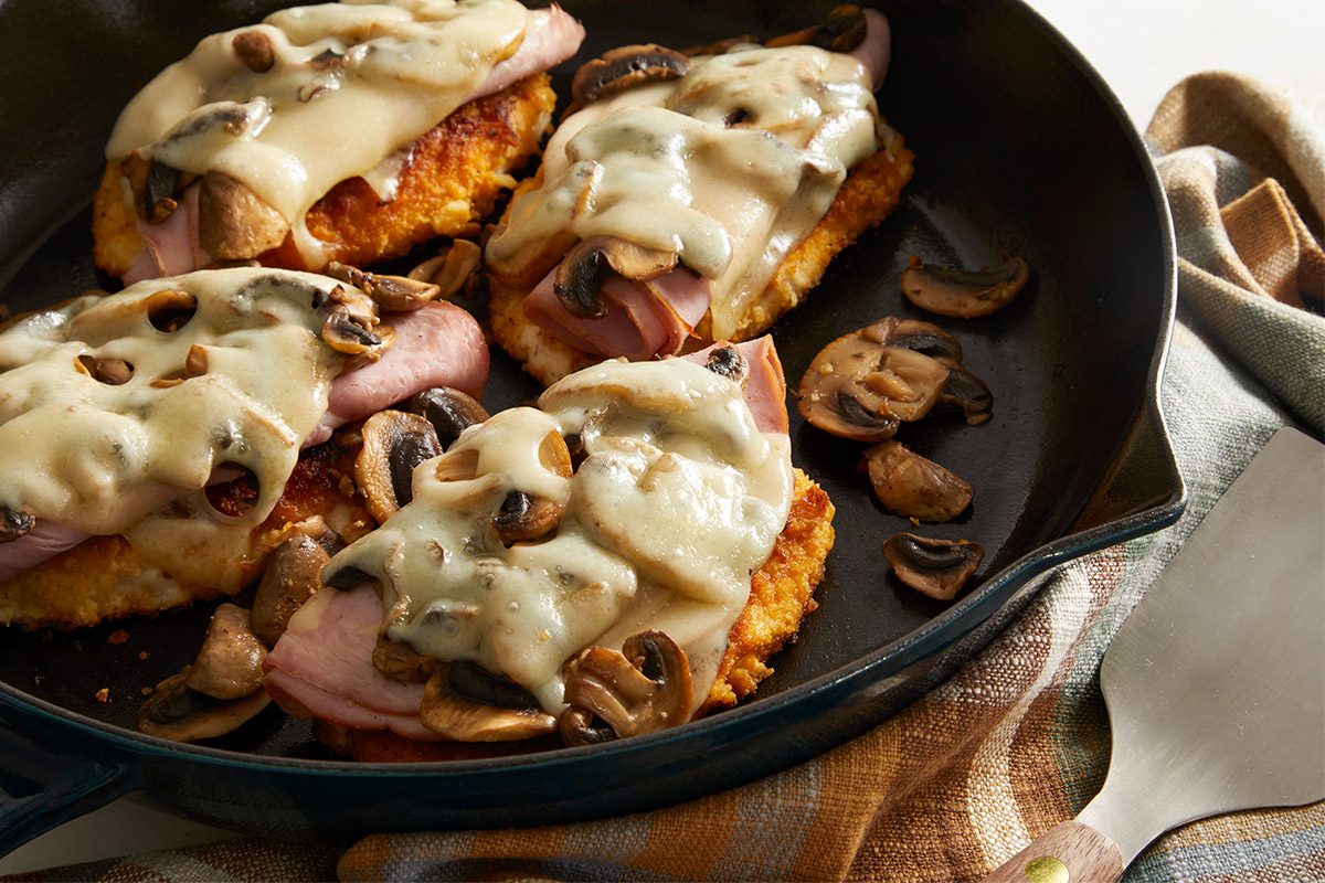 Close up shot of Swiss Mushroom Chicken topped with sautéed mushrooms, ham, and melted Swiss cheese in a black skillet on a plaid towel.