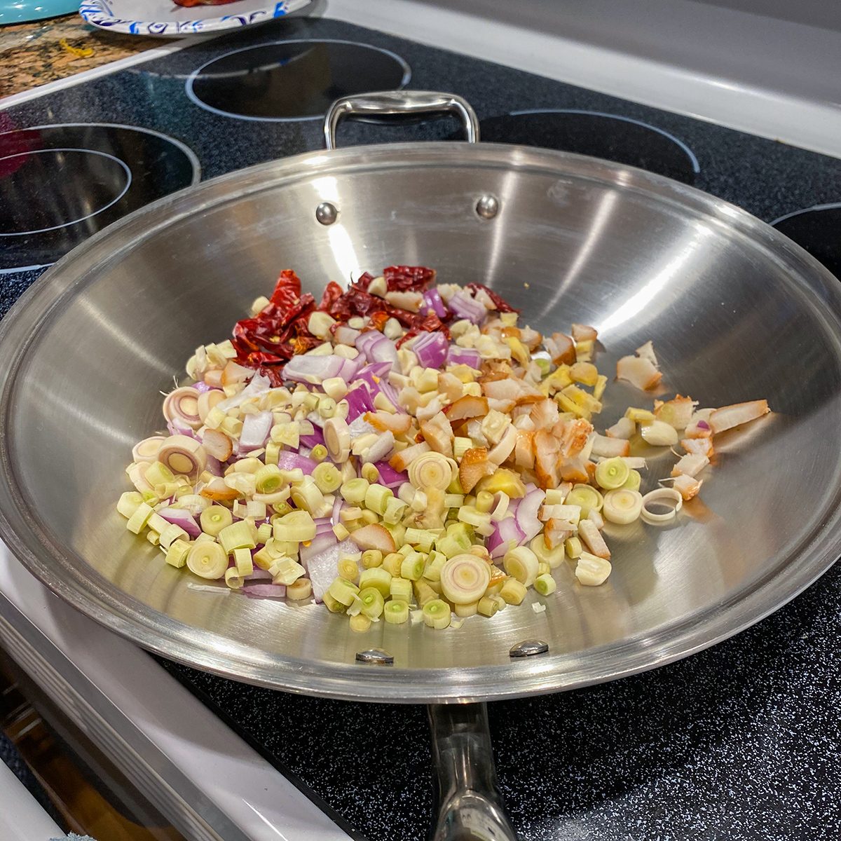 A stainless steel wok sits on a stovetop, filled with chopped vegetables including onions, lemongrass, and dried red chili peppers, ready to be cooked.