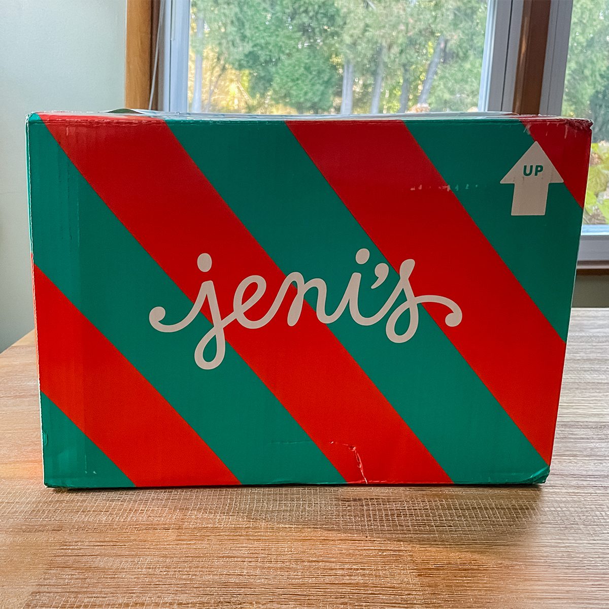 A box with red and green diagonal stripes and the word "Jeni's" written in white cursive sits on a wooden table in front of a window with a view of trees. An "up" arrow is printed in the top right corner of the box.
