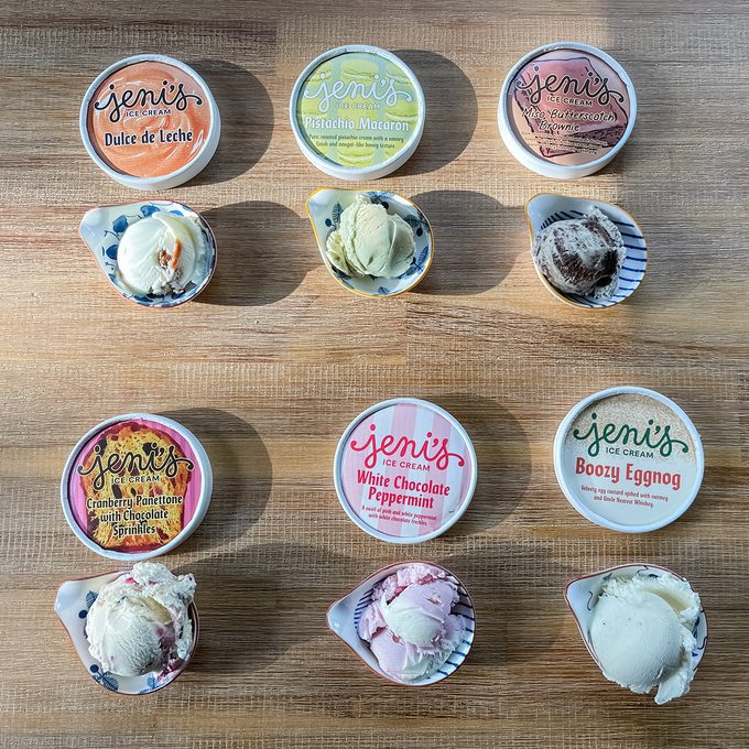 Nine scoops of ice cream on a wooden surface, each paired with a Jeni's Splendid Ice Creams container lid. Flavors include Dulce de Leche, Lemon, Dark Chocolate, Boozy Eggnog, White Chocolate Peppermint, and more.