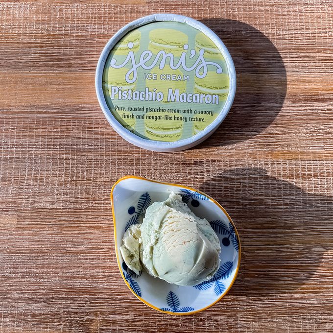 A small bowl with two scoops of pale green pistachio macaron ice cream sits beside a container labeled "Jeni's Ice Cream Pistachio Macaron" on a wooden surface.