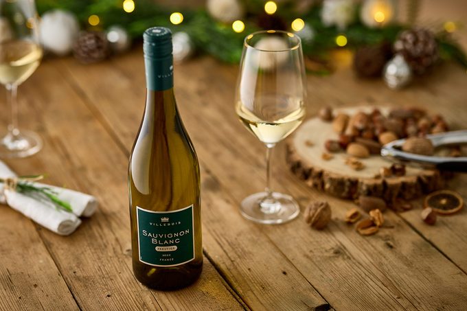 A bottle of Sauvignon Blanc wine stands next to a filled wine glass on a wooden table, decorated with nuts, a napkin, and festive evergreen garland and lights in the background.