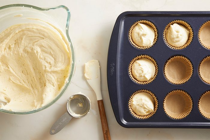A bowl of cupcake batter, a spatula, a metal scoop, and a muffin tin lined with paper baking cups, some filled with batter, are arranged on a light countertop.