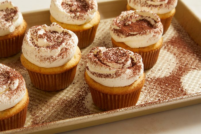 Vanilla cupcakes with swirled white frosting and cocoa powder dusting are arranged on a baking sheet, with extra cocoa powder sprinkled around them.