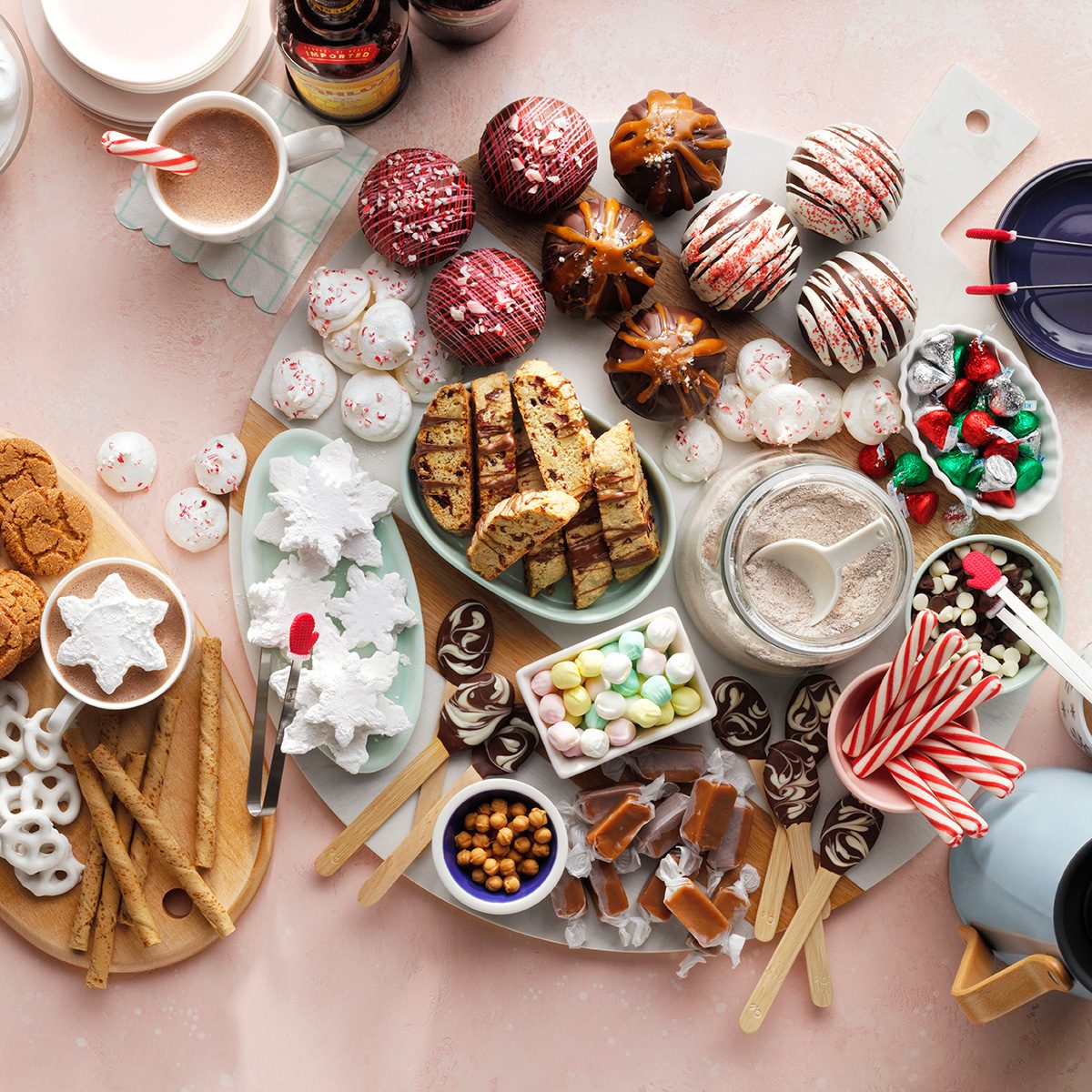 A festive assortment of holiday treats on a table, including cupcakes, cookies, biscotti, pretzels, candies, hot chocolate, and candy canes arranged on serving boards with mugs nearby.
