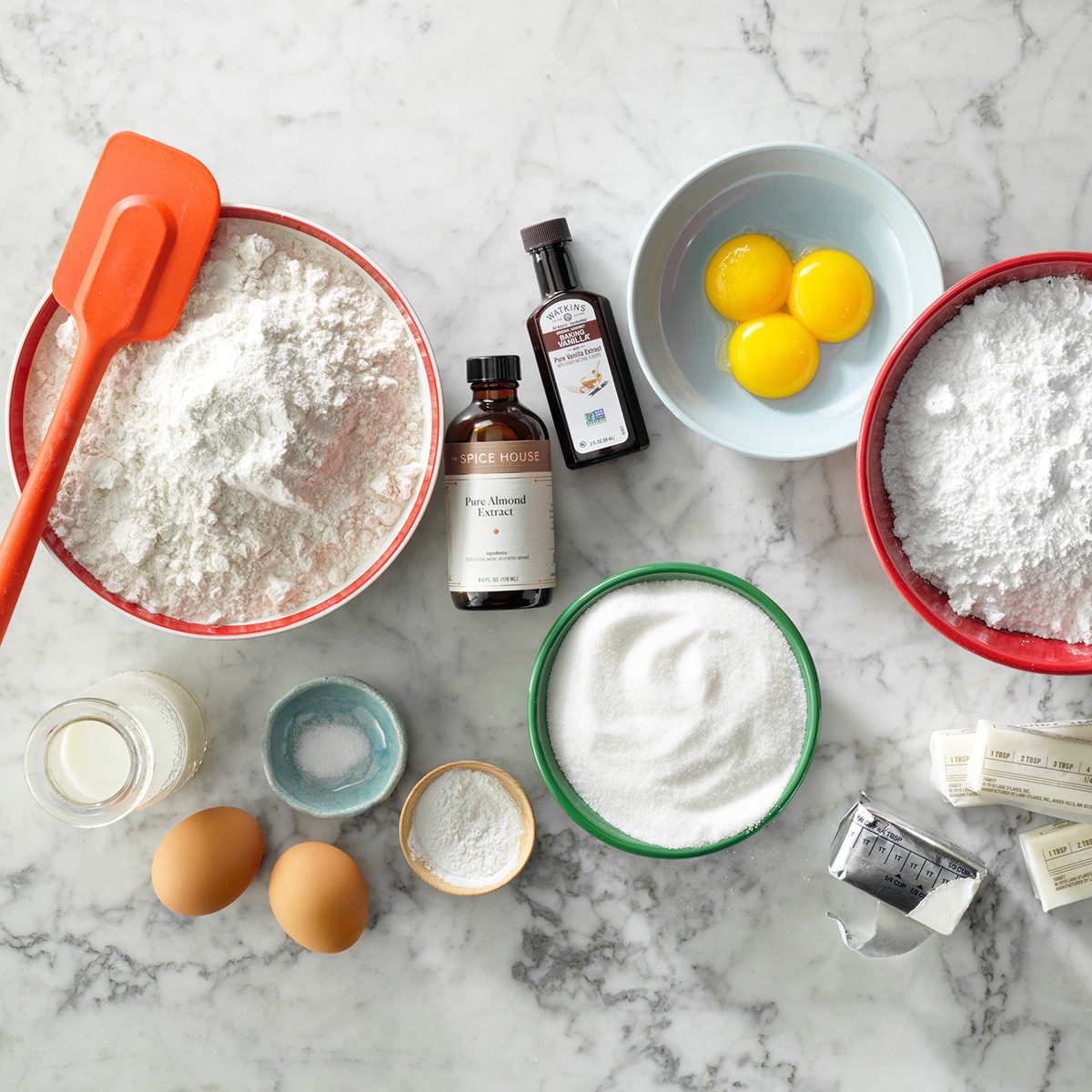 Baking ingredients on a marble surface: bowls of flour, sugar, powdered sugar, and eggs, with sticks of butter, small bottles of vanilla, a spatula, baking powder, milk, and extra eggs.