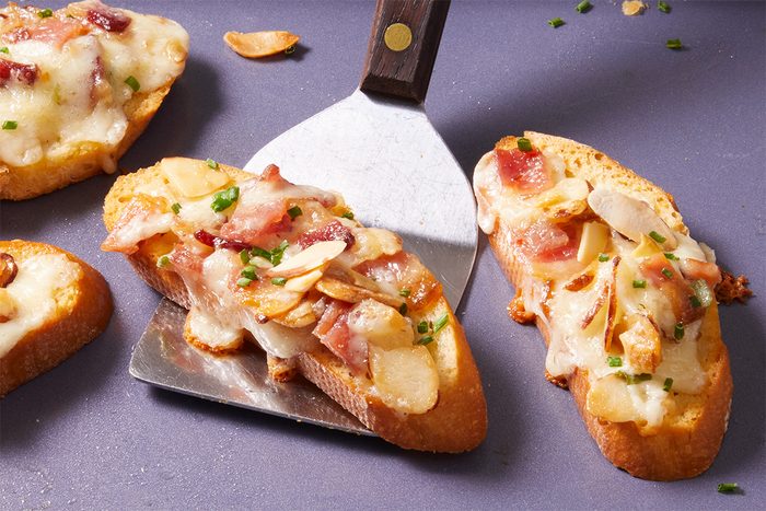 Close-up shot of Almond-Bacon Cheese Crostini pieces being lifted from a tray with a metal spatula, showing melted cheese and bacon bits on top.