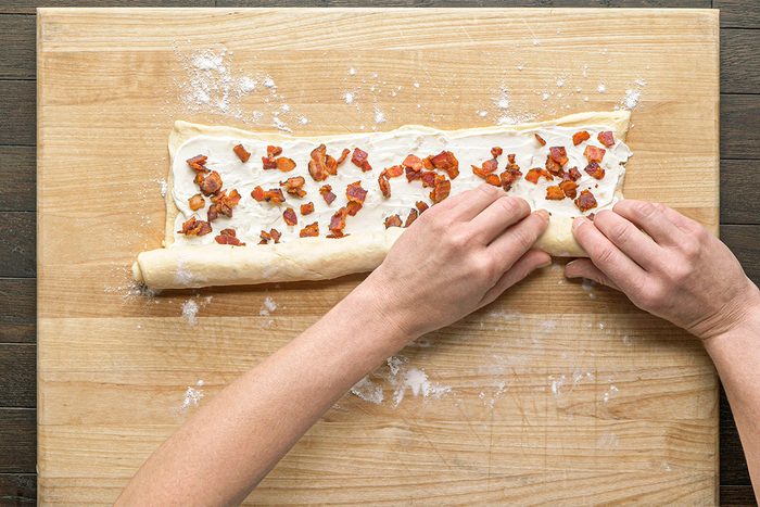 Close-up shot; a person rolls up dough spread with cream cheese; sprinkled bacon on floured wooden surface
