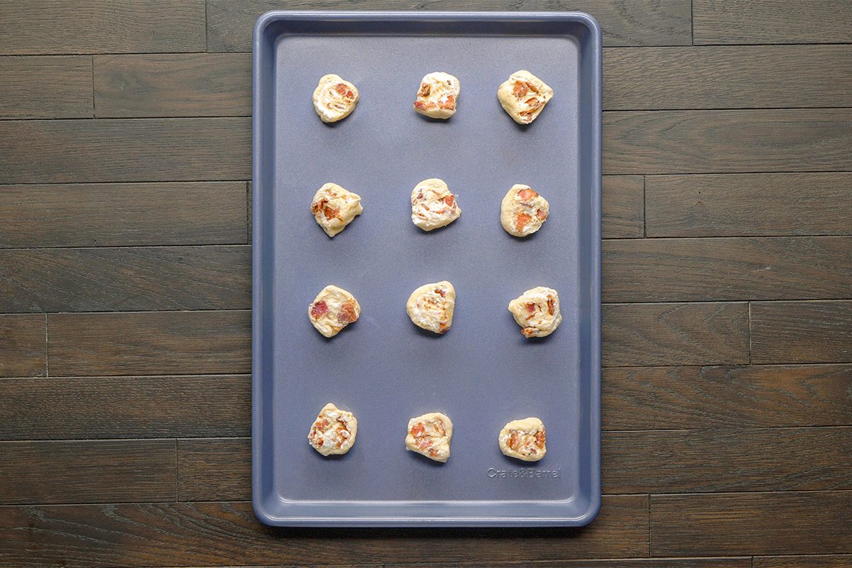 Overhead shot; a baking sheet holds 12 dough balls with meat and cheese bits on a dark wood surface