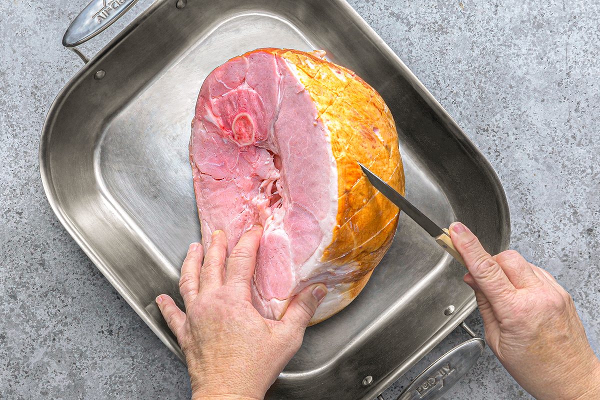 A person uses a knife to score the surface of a large, uncooked ham placed in a metal roasting pan on a gray countertop.