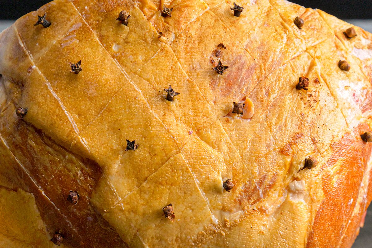 Close-up of a baked ham with a golden-brown surface, scored in a diamond pattern and studded with whole cloves at each intersection.