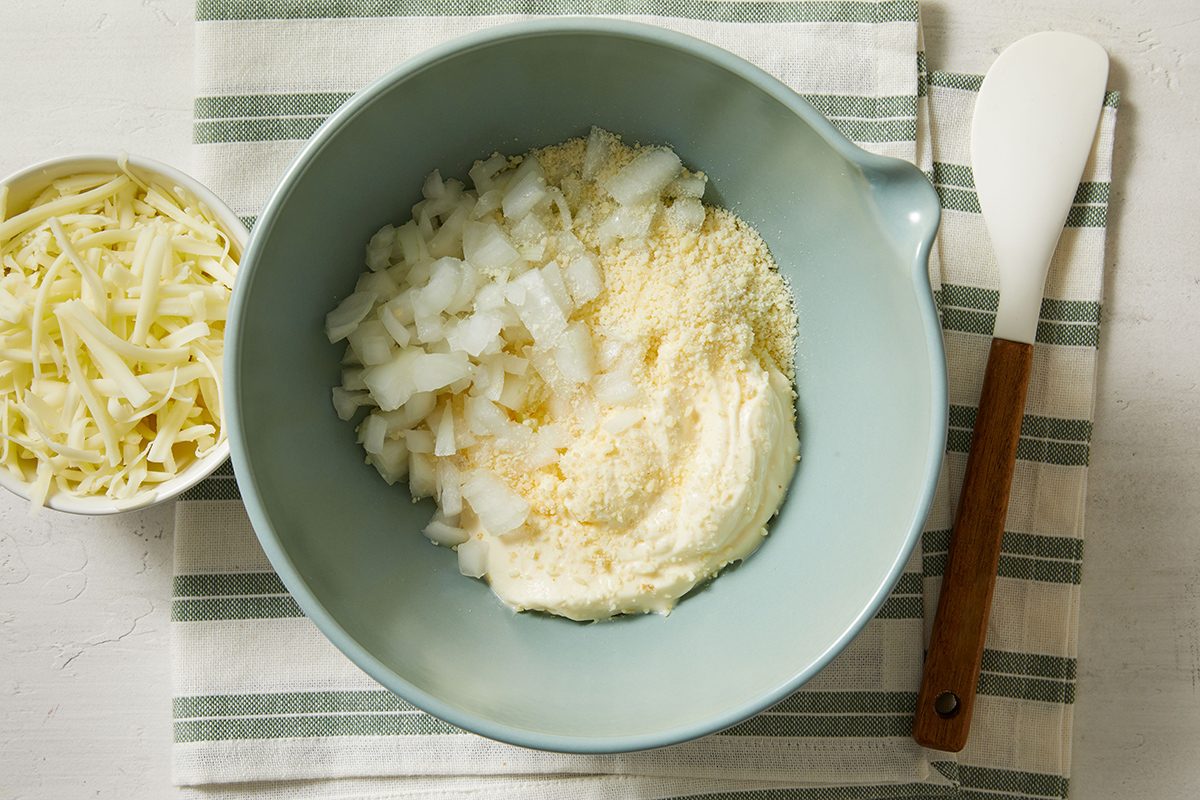 A blue mixing bowl with chopped onions, grated cheese, and ricotta cheese sits on a striped cloth next to a small bowl of shredded cheese and a white spatula with a wooden handle.
