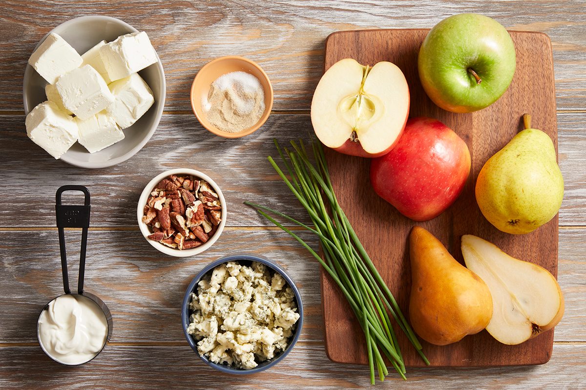 Overhead view of apples, pears, and chives on a wooden cutting board, surrounded by bowls of cream cheese, blue cheese, pecans, mayonnaise, and butter on a wooden surface.