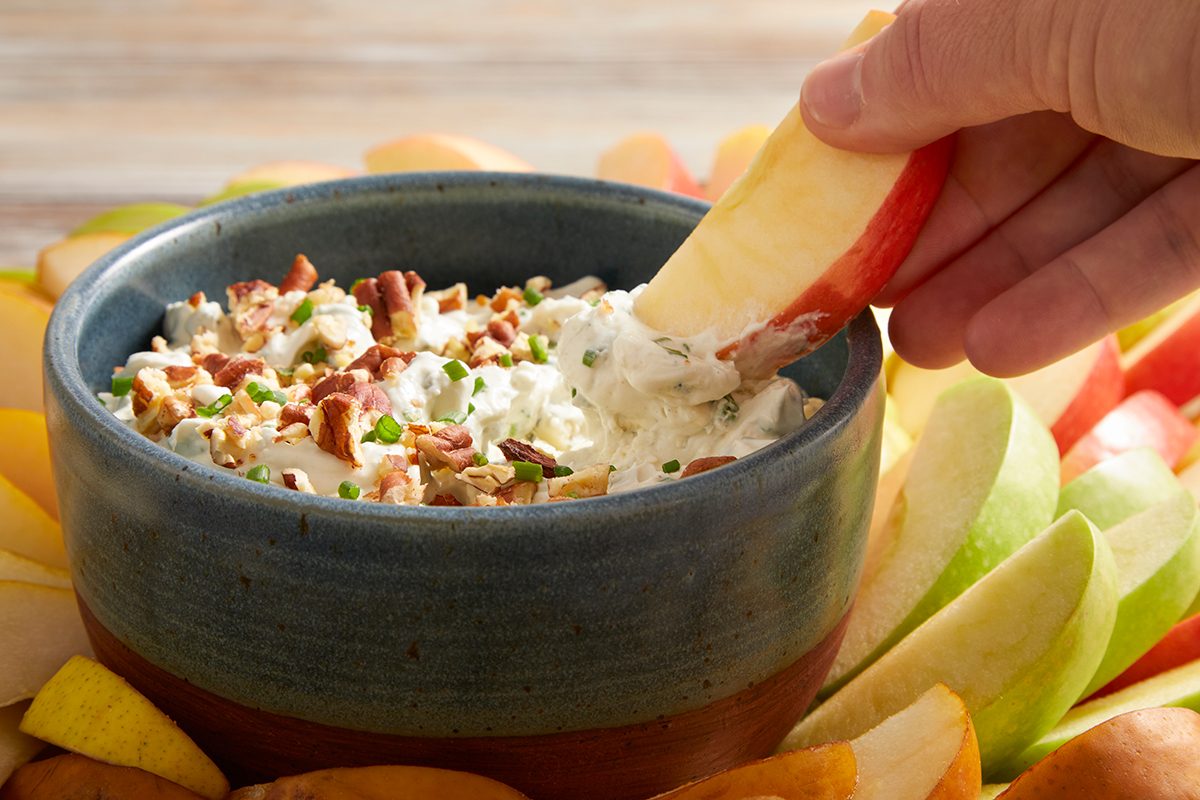 A hand dips an apple slice into a bowl of creamy dip topped with chopped pecans and chives, surrounded by assorted apple slices.