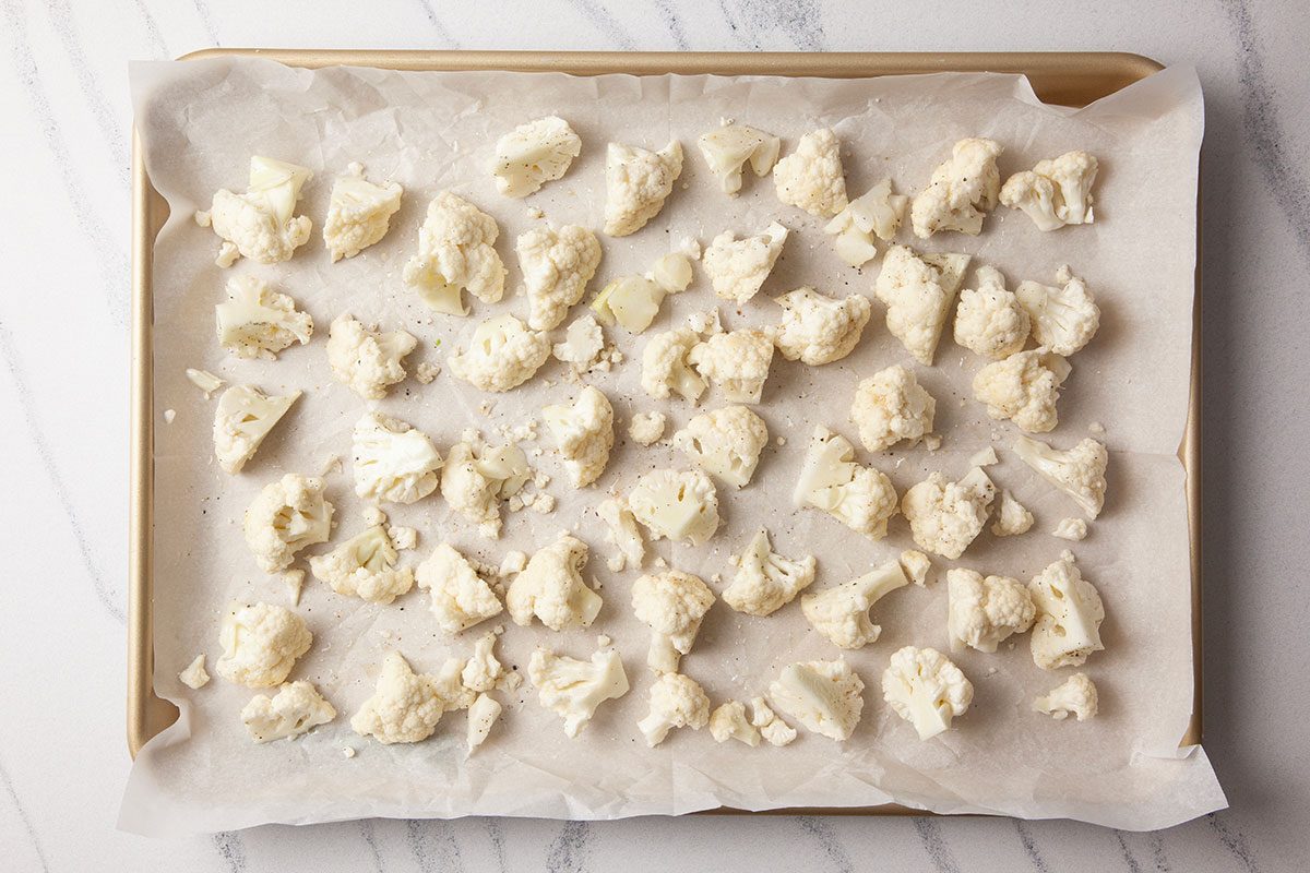 Overhead shot of raw cauliflower florets spread evenly on a parchment-lined baking sheet, set on a light stone surface;