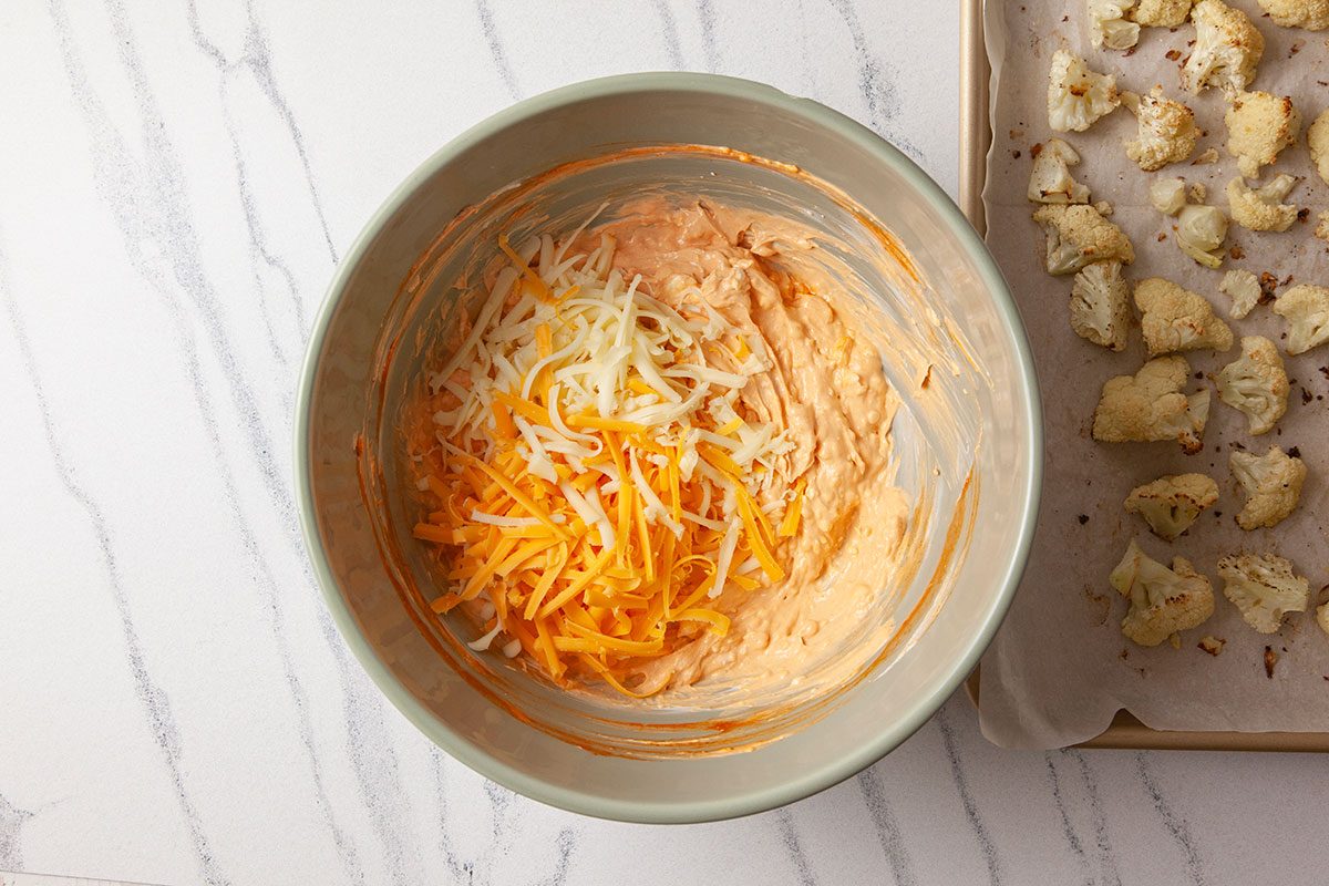 Overhead shot of a bowl of creamy orange mixture topped with shredded cheese, set beside a parchment-lined tray of roasted cauliflower on a light marble surface;