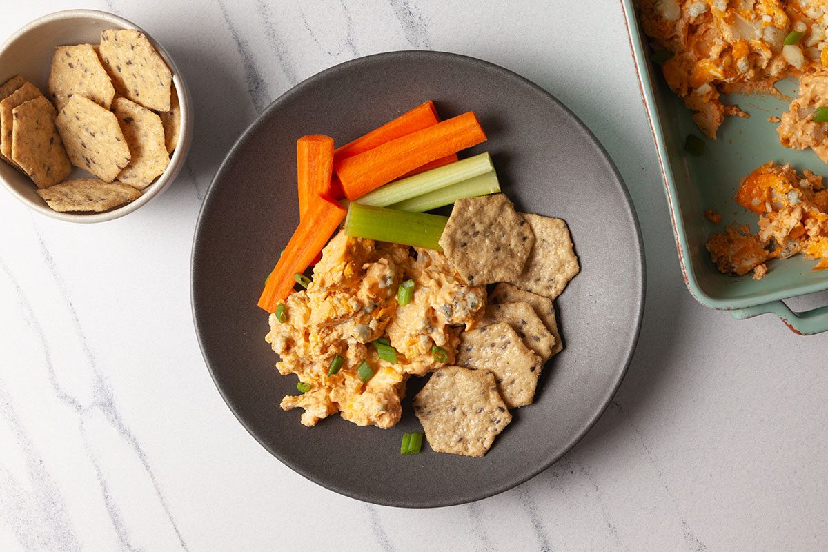 Overhead shot of creamy cauliflower dip served on a dark gray plate with crackers, carrot and celery sticks, set on a light marble surface;