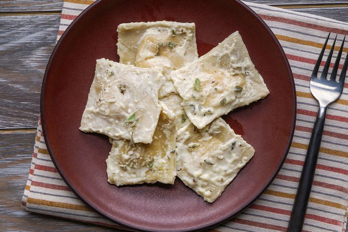 Overhead shot of Butternut Squash Ravioli covered in creamy white sauce and herbs rests on a striped cloth napkin with a fork to the right of the plate