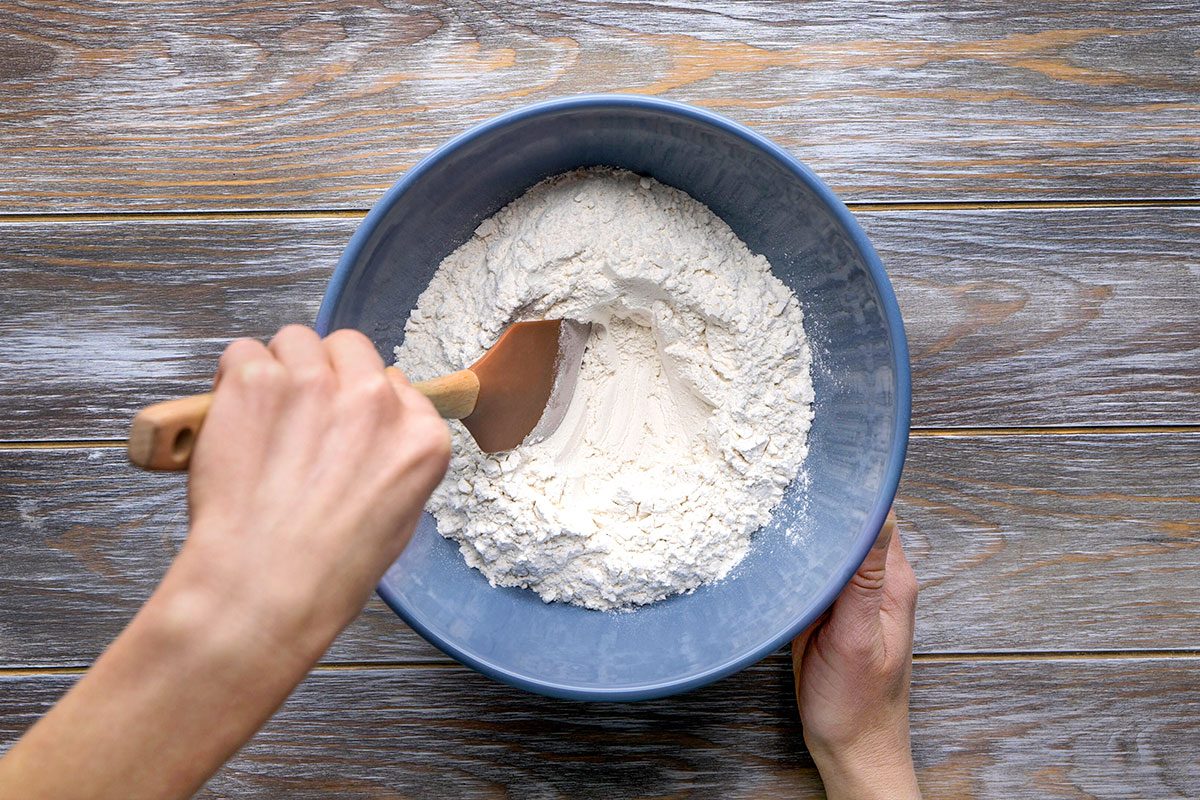Overhead shot of a person stirs white flour in a blue bowl using a wooden spatula on a wooden surface and the ingredients are being mixed gently