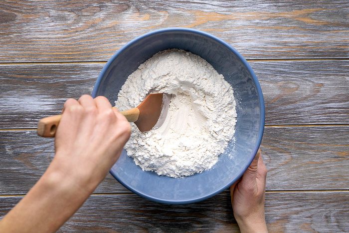 Overhead shot of a person stirs white flour in a blue bowl using a wooden spatula on a wooden surface and the ingredients are being mixed gently