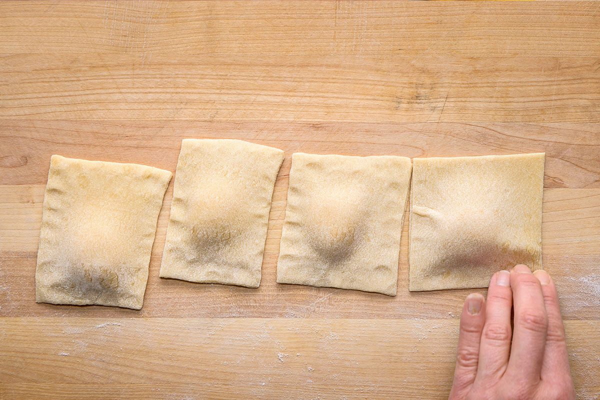 Overhead shot of a four uncooked ravioli squares are arranged on a floured wooden surface and a hand is gently pressing the edge of the last ravioli