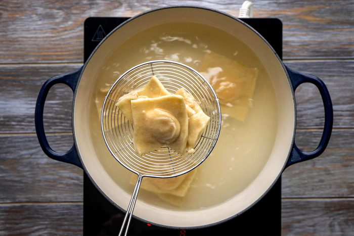 Overhead shot of a metal strainer lifts ravioli from a pot of boiling water on a stovetop and the wooden surface is in the background nearby