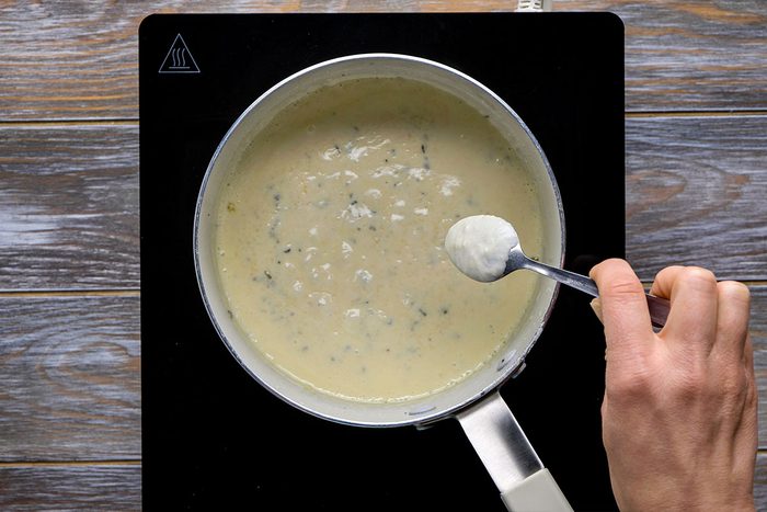 Overhead shot of a hand holds a spoon over a saucepan filled with creamy white herb sauce it is heating on a black stovetop placed on wooden surface