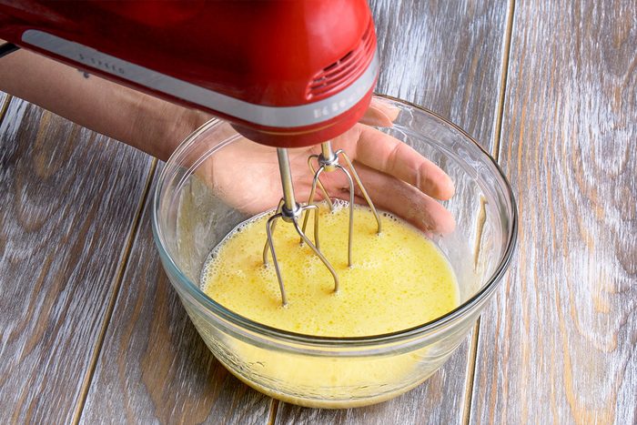 3/4 angle view shot of a person is using a red electric hand mixer blending a yellow liquid in a clear glass bowl placed on a wooden surface likely eggs