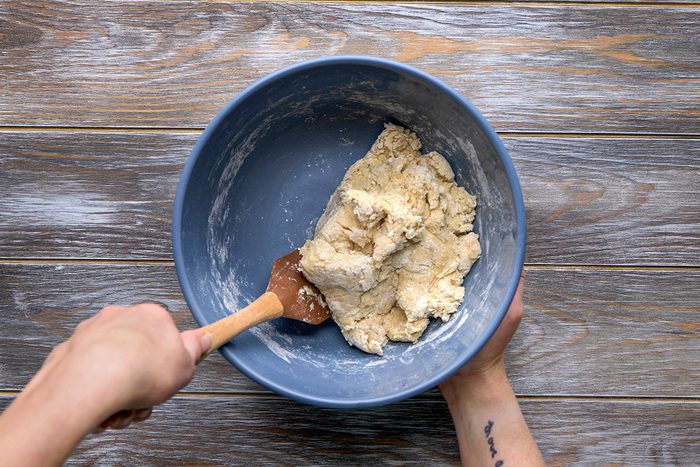 Overhead shot of a person mixes dough with a spatula in a large blue bowl and the bowl sits on a wooden surface as the mixing continues