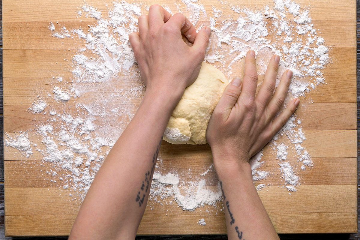Overhead shot of a person kneads dough on a floured wooden surface and uses both hands while flour covers the board and tattoos show on their forearms