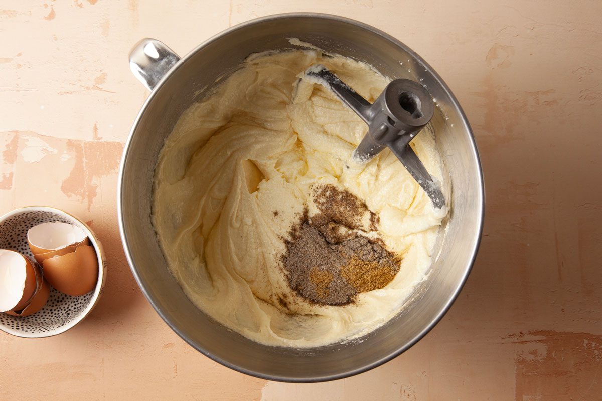 Overhead shot of a mixing bowl holds creamy batter with spices and a paddle attachment next to it a small bowl has two cracked eggshells