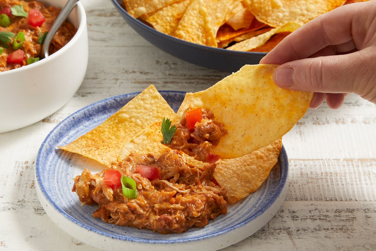A hand dips a tortilla chip topped with shredded chicken, tomato, and herbs from a blue-rimmed plate; a bowl of chips and a bowl of chicken are in the background.