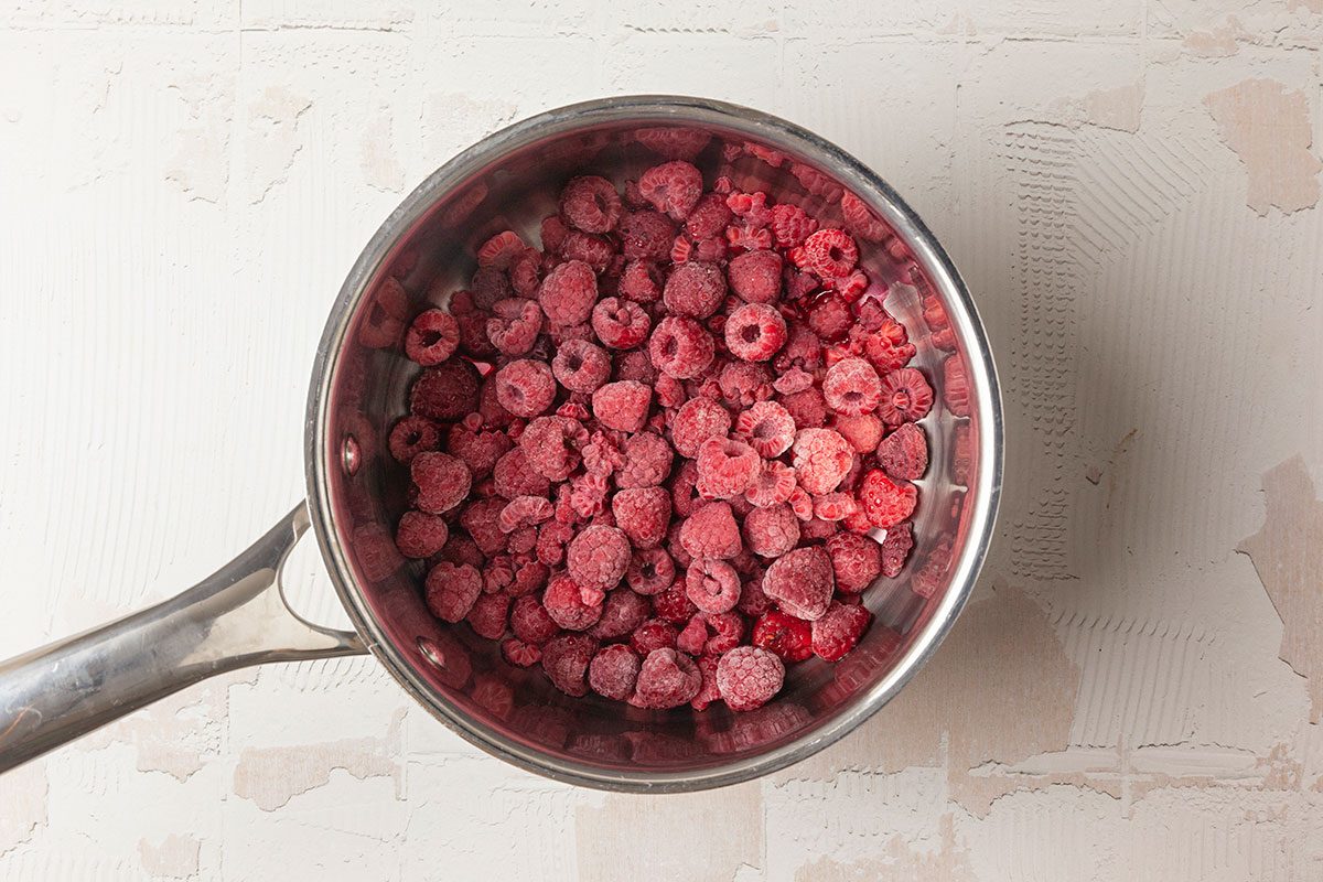 Overhead shot of frozen raspberries piled in a saucepan, ready for cooking, set on a light textured surface;