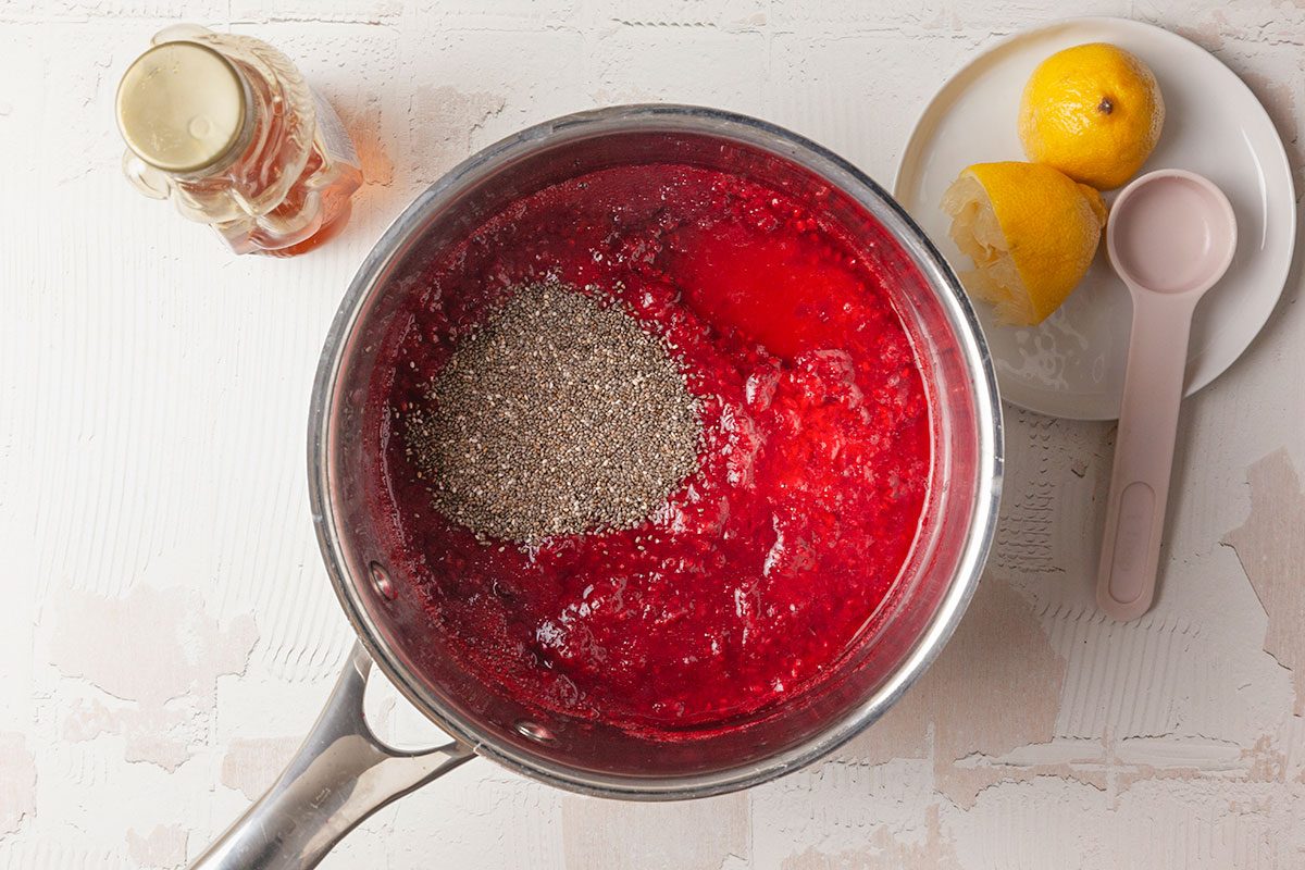 Overhead shot of simmered raspberries topped with chia seeds in a saucepan, with lemon and sweetener nearby on a light textured surface;