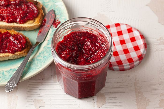 3/4 angle shot of a clear glass jar filled with glossy red chia seed jam with visible fruit and seeds, set on a light textured surface with jam-covered toast softly in the background;