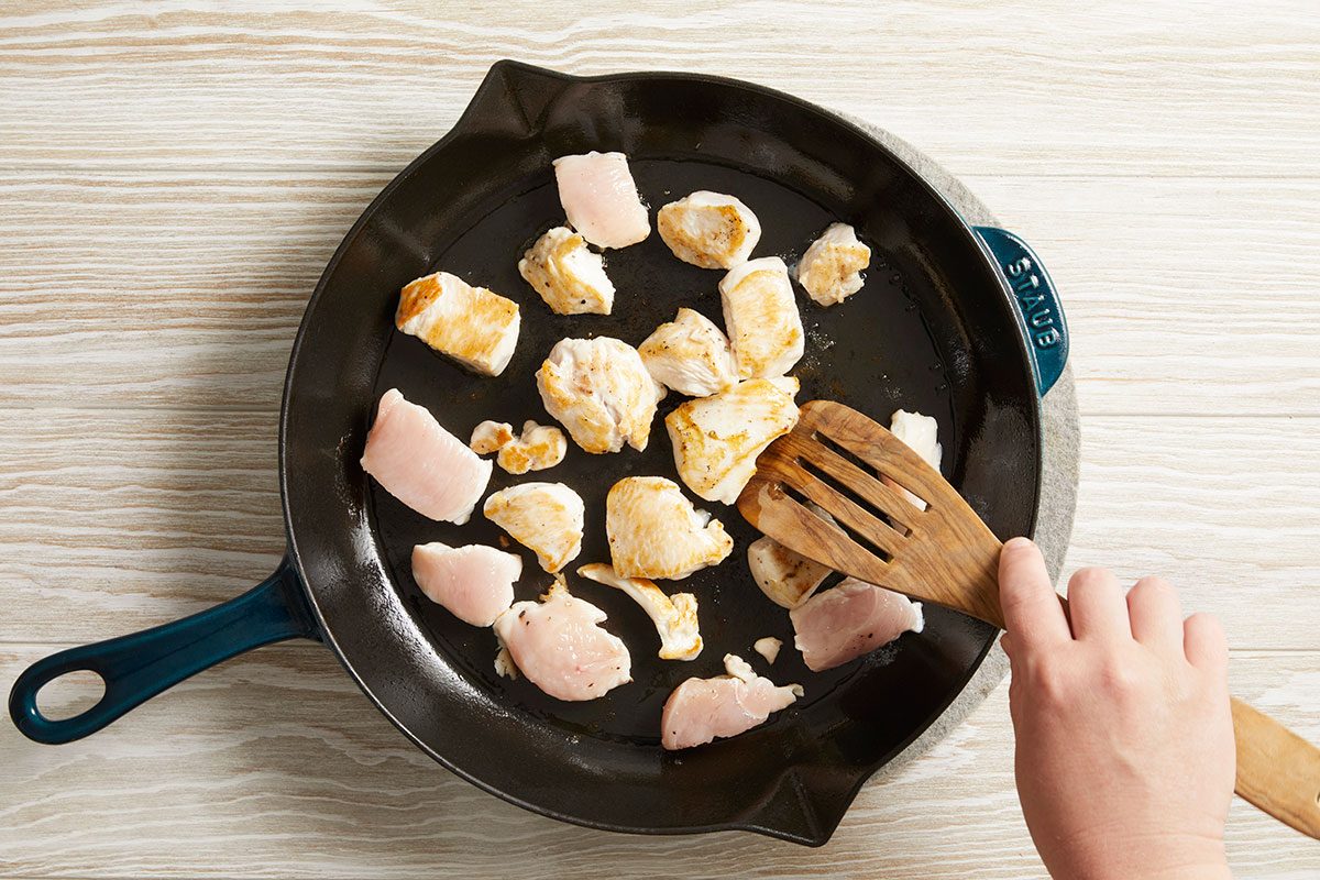 Overhead shot of chicken pieces cooking in a skillet, hand stirring with spatula.