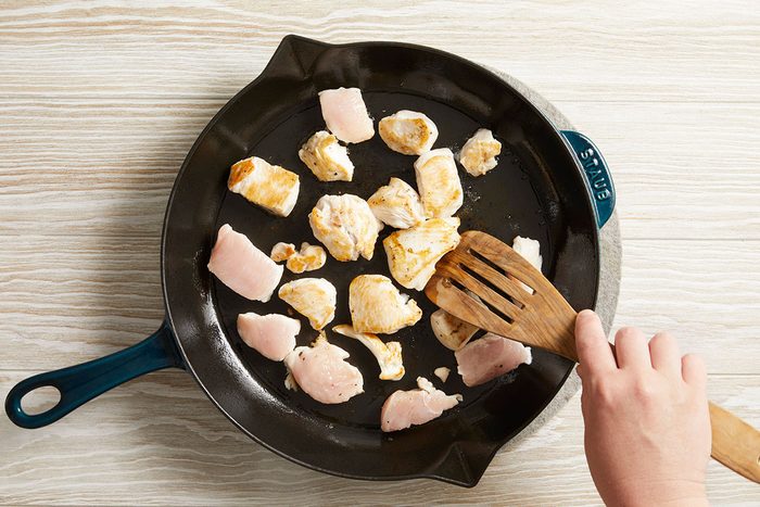 Overhead shot of chicken pieces cooking in a skillet, hand stirring with spatula.