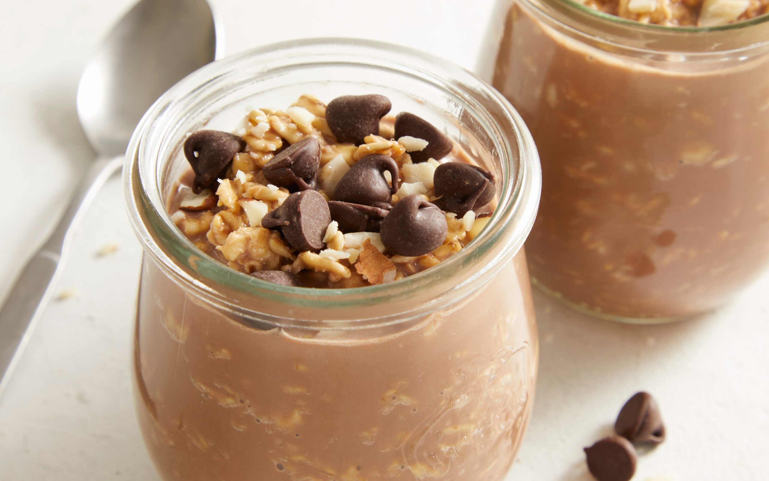 Close-up overhead shot of a glass jar filled with Chocolate Overnight Oats, topped with granola and chocolate chips, with another jar blurred in the background.