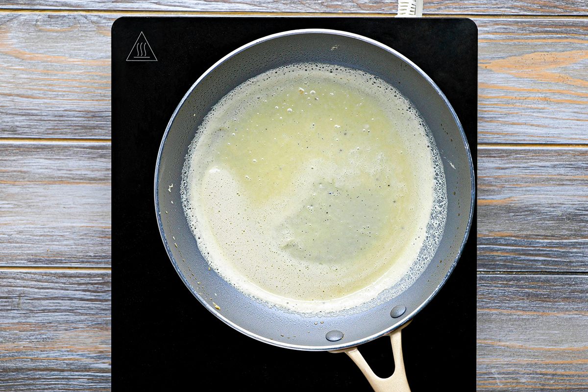 A frying pan with melted butter sits on a black stovetop over a wood-patterned surface. The butter is foaming and partially covers the pan's bottom.