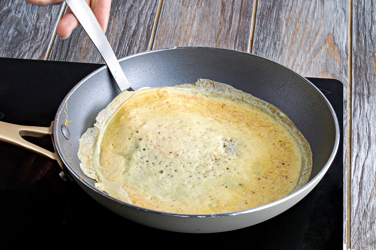 A hand uses a spatula to lift the edge of a thin crepe cooking in a nonstick frying pan on a stovetop with a wood-textured background.