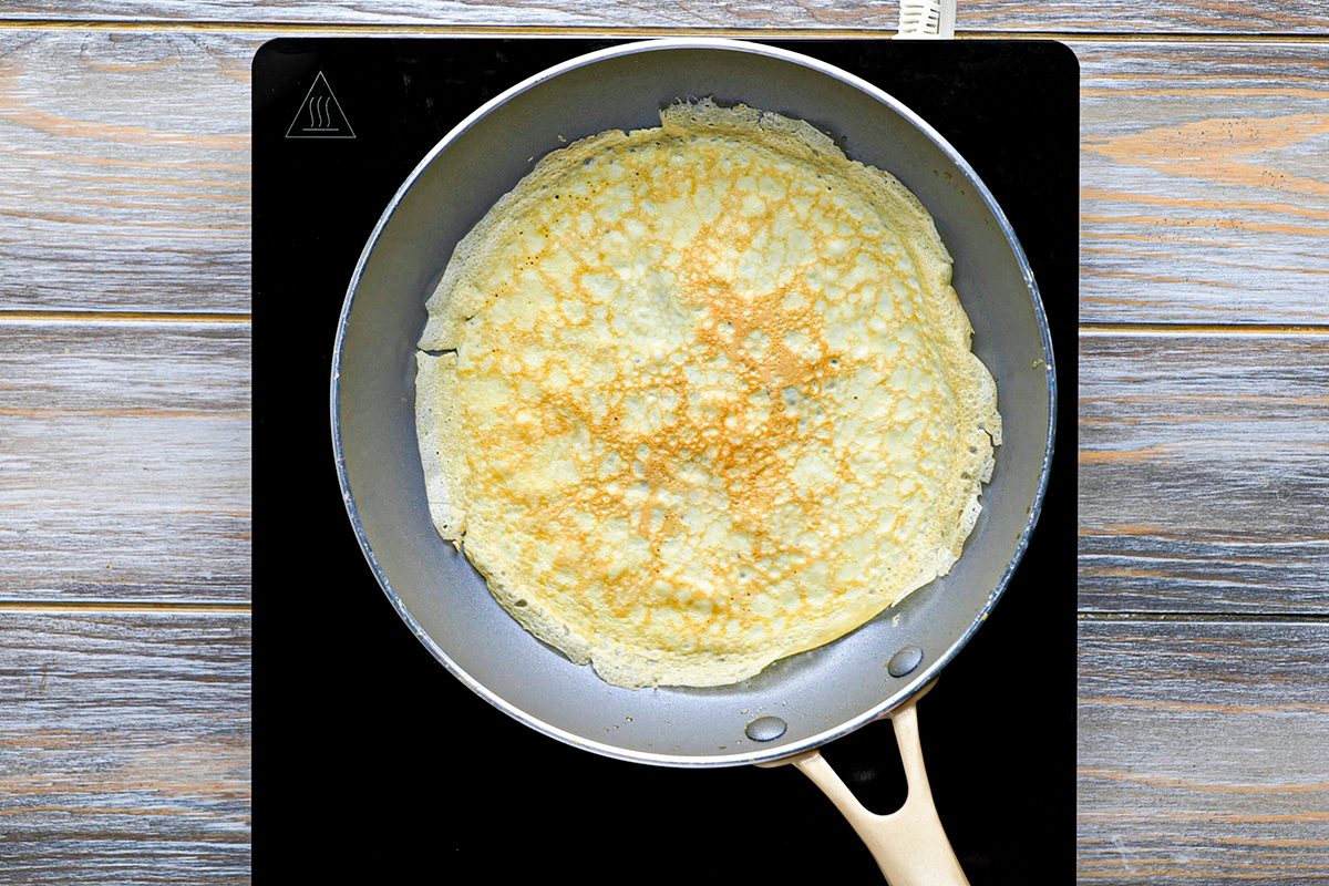A thin, golden crepe cooking in a frying pan on a black stovetop, viewed from above on a wooden surface.