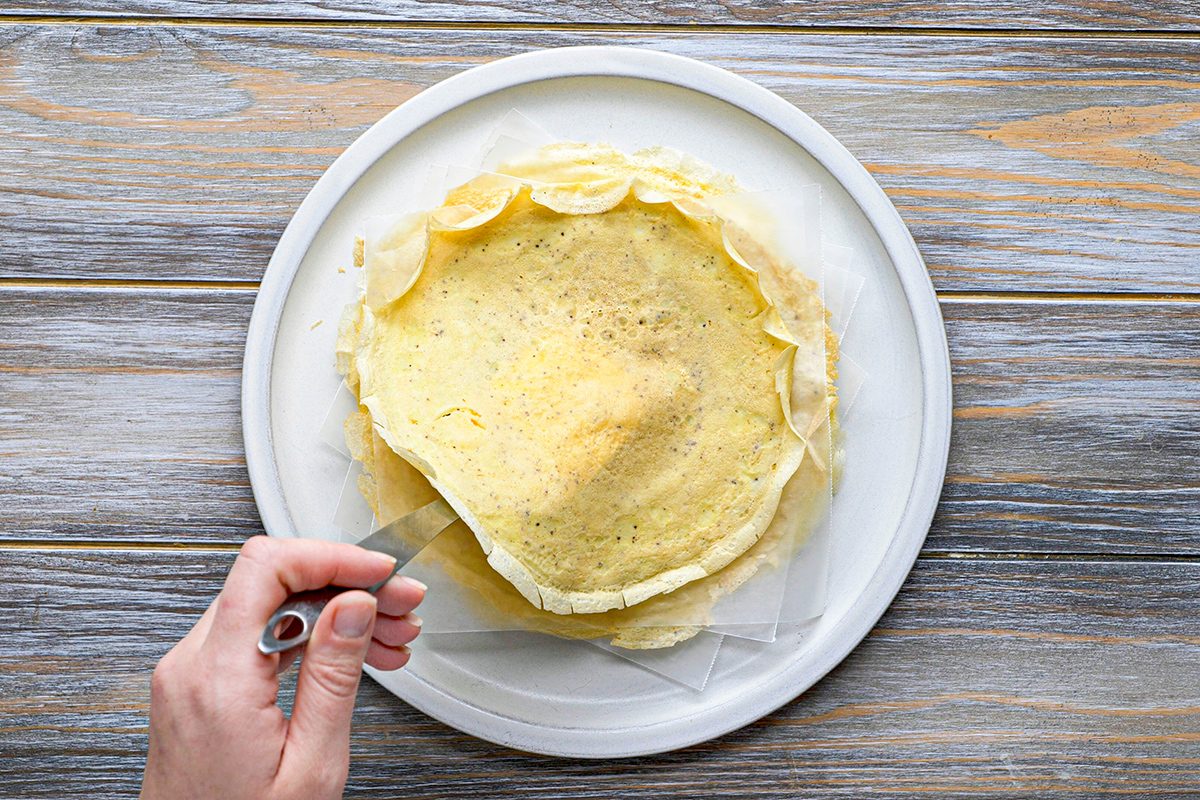 A hand lifts the edge of a thin crepe with a spatula from a stack of crepes on a white plate, set on a wooden surface.