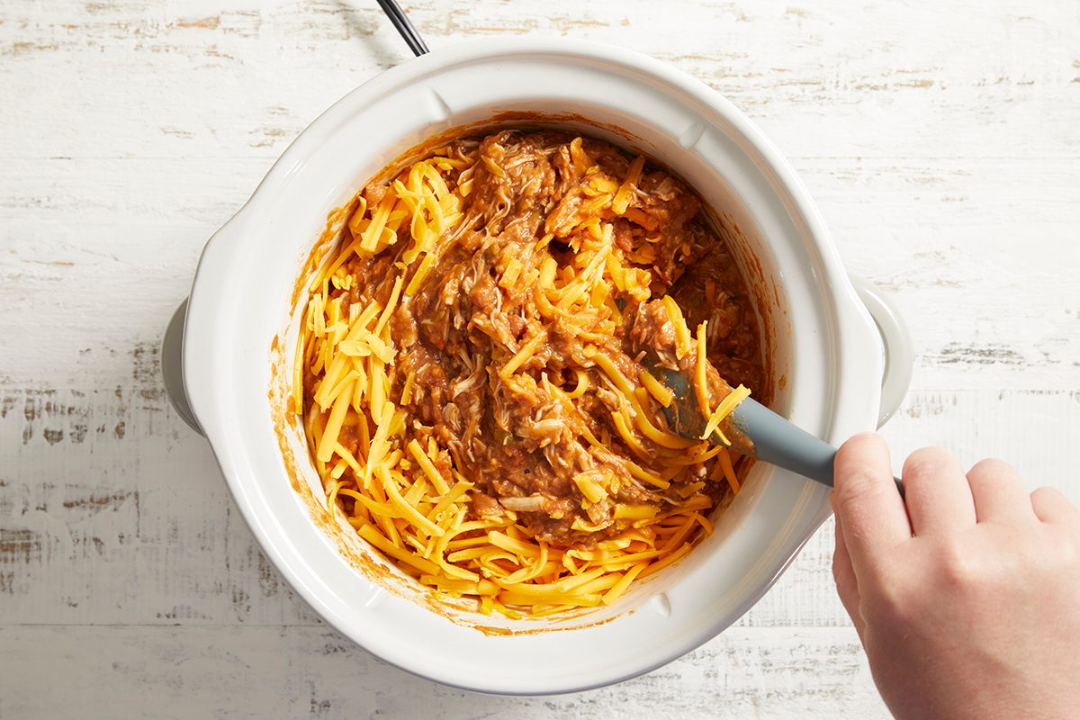 A hand stirs shredded cheese and brown seasoned meat in a white slow cooker on a light wooden surface.